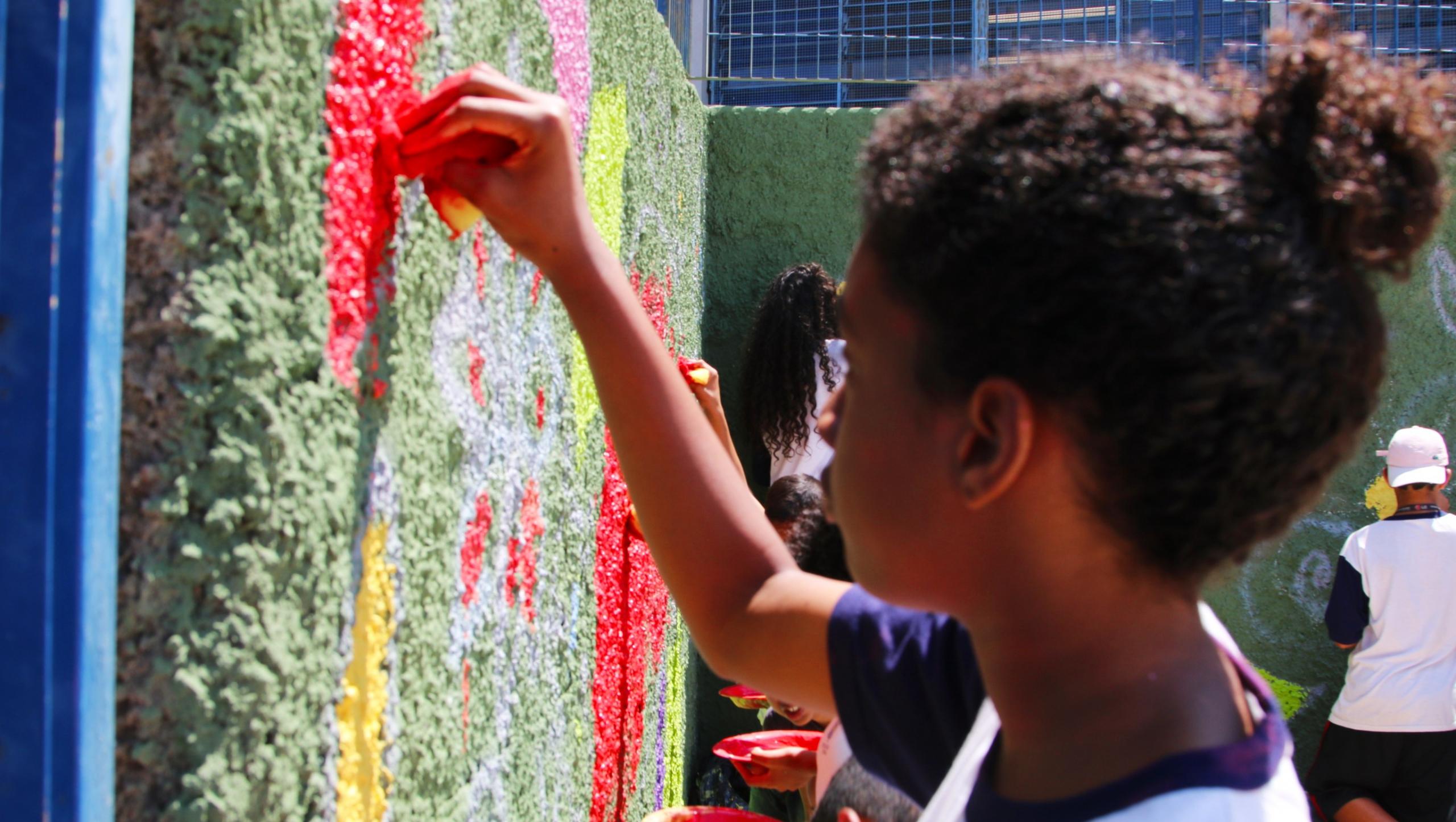 Jovens fazem desenhos abstratos em um muro verde. No primeiro plano, uma menina usa uma esponja para fazer um desenho vermelho.