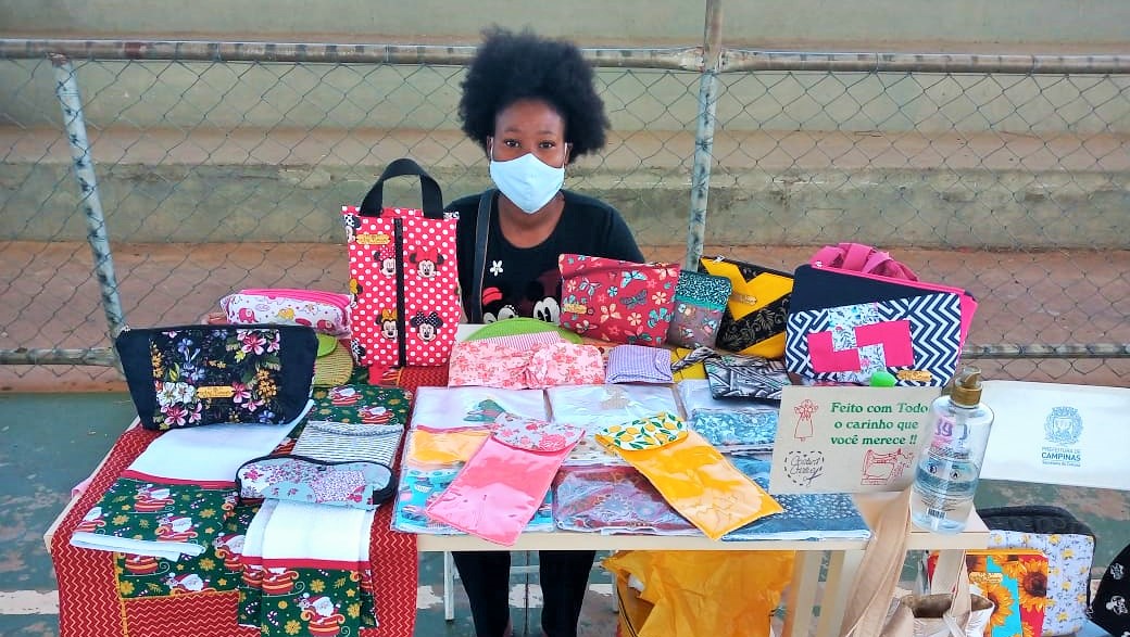 Mulher negra de máscara está sentada atrás de uma mesa mostrando uma série de produtos, como bolsas, panos de prato e sacolas, feitos com estampas bem coloridas