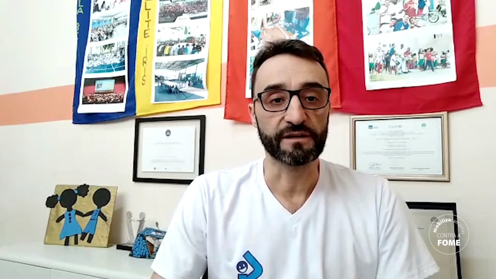 Claudio Raizaro, do Progen, de camiseta branca, óculos e barba, fala ohando para a câmera. trás, se veem quadros e uma prateleira com objetos