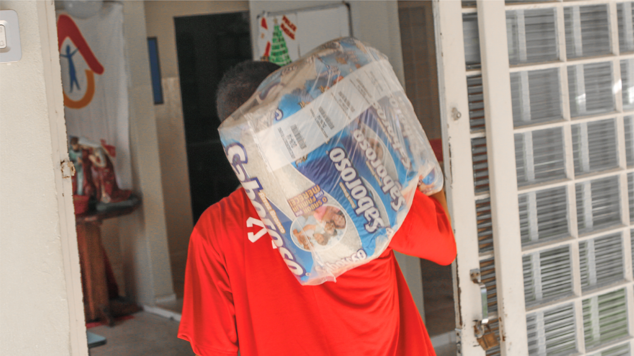Imagem mostra homem de costas, carregando uma cesta básica.