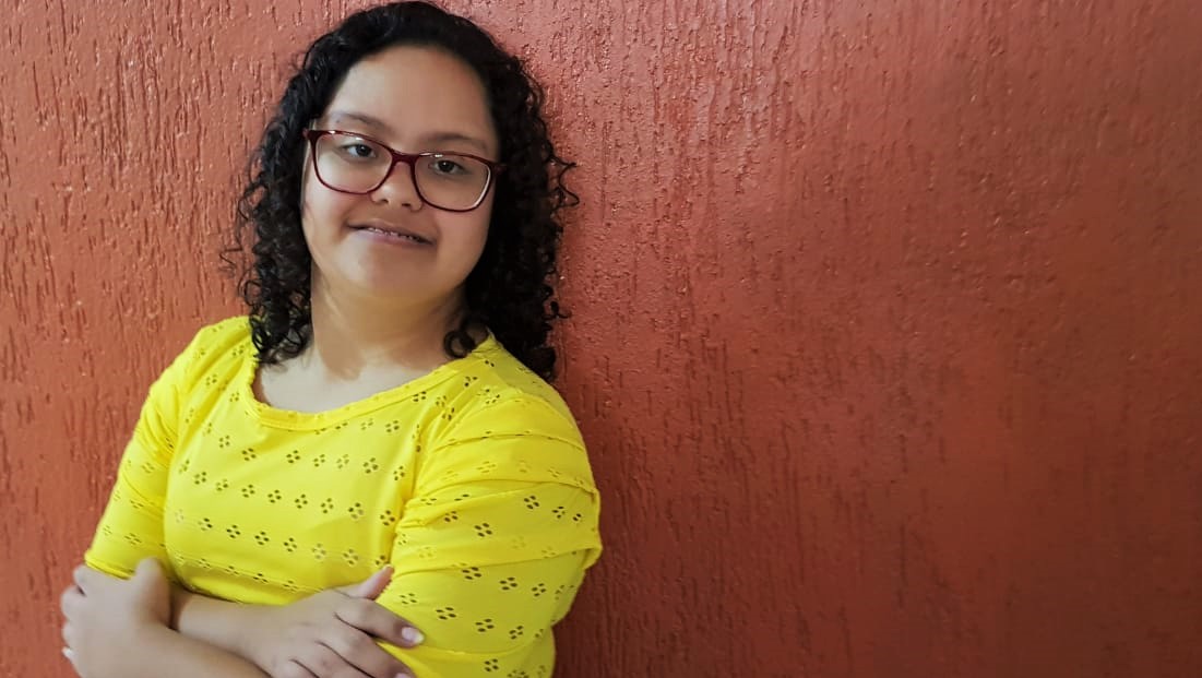Jovem com síndrome de Down, cabelos escuros cacheados e óculos, posa de braços cruzados, usando uma blusa amarela.