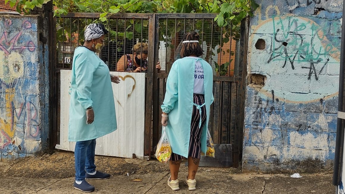 Imagem mostra três mulheres: uma está atrás do portão de uma casa; as outras duas estão com equipamentos de proteção contra a pandemia e segura seguras com alimentos