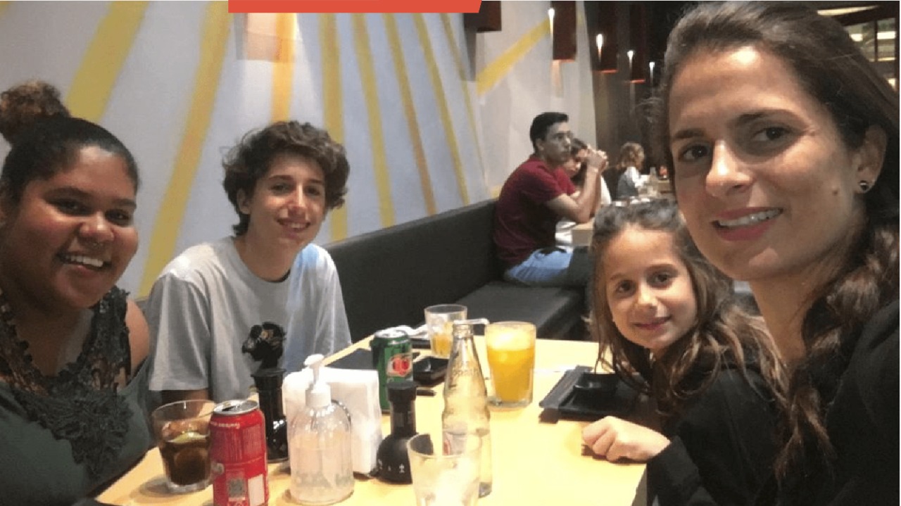 Uma mulher negra, duas crianças e uma mulher branca estão sentados numa mesa de restaurante, olhando diretamente para a câmera, sorrindo.