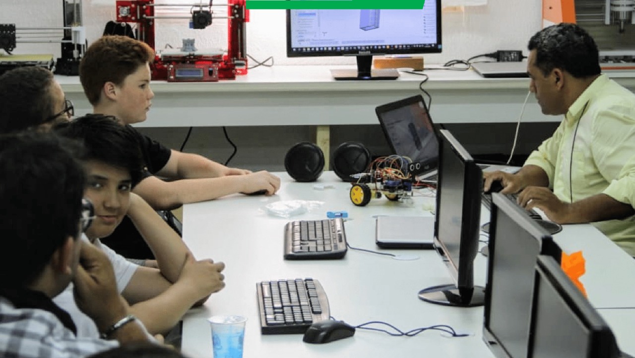 Em uma mesa grande, de um lado, estão jovens sentados na frente de computadores e do outro, uma pessoa mais velha, também na frente de um computador. Ao fundo, alguns equipamentos, como uma impressora 3-D