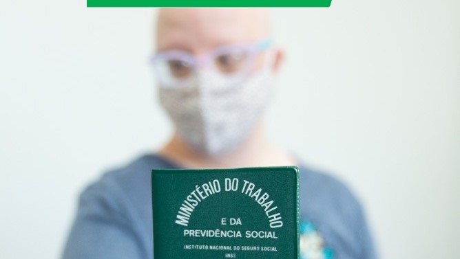 Foto mostra uma pessoa branca em segundo plano e desfocada, apontando uma carteira de trabalho para a câmera.