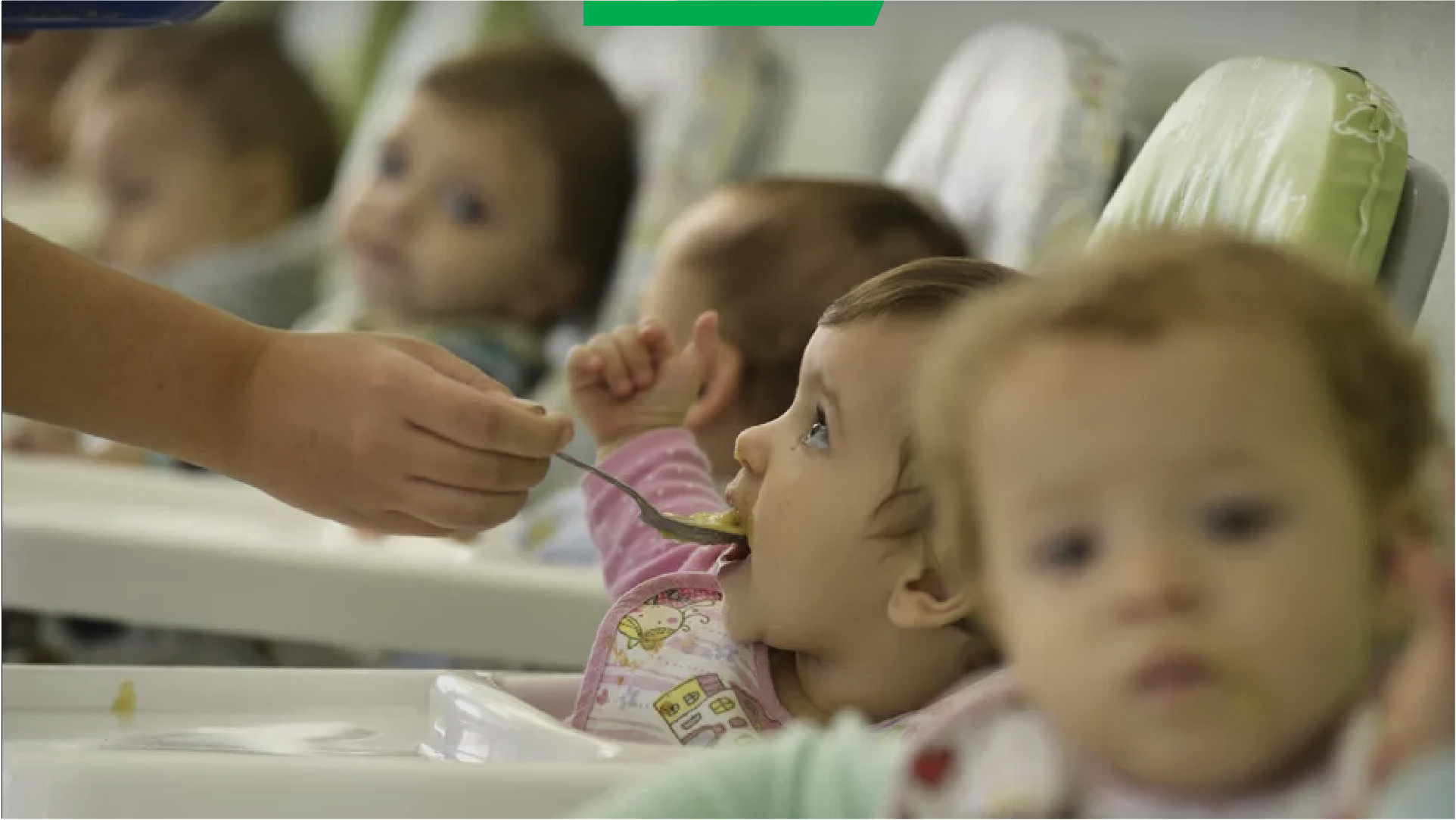 Bebê, sentada na cadeira de refeição, recebe uma colher de alimento